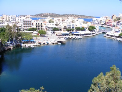 A scenic view of Agios Nikolaos harbor in Crete, Greece, with calm blue waters surrounded by cafes, shops, and buildings. The harbor opens out to the sea, with a small island visible in the distance, showcasing the vibrant coastal town and its relaxed charm. A scenic view of Agios Nikolaos harbor in Crete, Greece, with calm blue waters surrounded by cafes, shops, and buildings. The harbor opens out to the sea, with a small island visible in the distance, showcasing the vibrant coastal town and its relaxed charm.