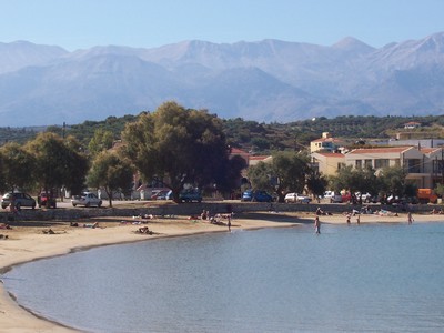 A scenic view of Almyrida Beach in Crete, Greece, with calm turquoise waters, sunbathers on the sandy shore, and the majestic White Mountains in the background.