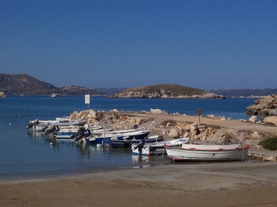 A picturesque view of Almyrida Harbour in Crete, Greece, featuring small fishing boats moored along the shoreline with a backdrop of rocky islets and the deep blue sea.