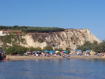 A scenic view of Almyrida Beach, Crete, with golden sand, sun loungers, and umbrellas set against a dramatic white cliff backdrop and lush greenery.