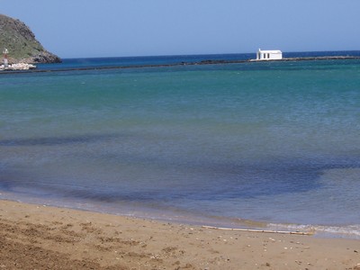 Sandy beach at Georgiopolis, Crete, with clear turquoise waters and a small white chapel on a rocky outcrop in the distance, typical of the serene coastal landscapes in Crete.