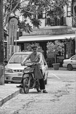 Black and white photo of a man sitting on a motorbike in front of parked cars in a quaint street, Chania, evoking a rustic charm.