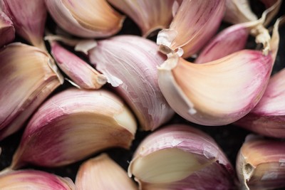 A close-up of fresh garlic cloves with their papery skins partially peeled, revealing a mix of creamy white and purple hues. The cloves are clustered together, showcasing their natural texture and rich color, essential for Mediterranean and Cretan cuisine.