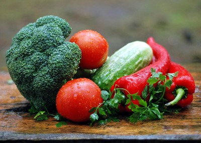 A fresh assortment of vegetables including broccoli, ripe red tomatoes, a cucumber, and vibrant red chili peppers, garnished with sprigs of parsley. The vegetables are covered in tiny water droplets and placed on a rustic wooden surface, highlighting their freshness and natural appeal.
