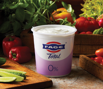 A tub of FAGE Total 0% Greek yogurt placed on a rustic kitchen counter, surrounded by fresh vegetables including cucumbers, tomatoes, and peppers. The creamy texture of the yogurt is visible, emphasizing its rich and smooth consistency, perfect for healthy Mediterranean-inspired meals.