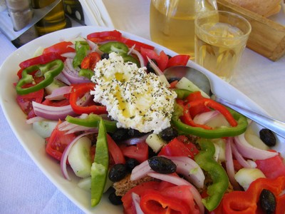 A colourful plate of traditional Cretan salad with fresh tomatoes, cucumbers, red onions, green and red bell peppers, olives, and a generous topping of creamy Mizithra cheese sprinkled with herbs, served alongside a glass of white wine. A colourful plate of traditional Cretan salad with fresh tomatoes, cucumbers, red onions, green and red bell peppers, olives, and a generous topping of creamy Mizithra cheese sprinkled with herbs, served alongside a glass of white wine.