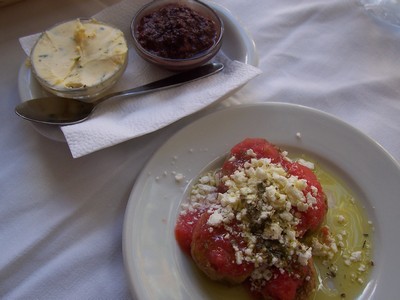 A plate of traditional Cretan dakos, featuring a barley rusk topped with chopped ripe tomatoes, crumbled feta or mizithra cheese, and a generous drizzle of olive oil, garnished with herbs. Beside it, there are two small bowls—one containing a creamy herb butter and the other a dark olive tapenade—served with a spoon on a white napkin. The simple yet flavorful presentation highlights classic Mediterranean ingredients. A plate of traditional Cretan dakos, featuring a barley rusk topped with chopped ripe tomatoes, crumbled feta or mizithra cheese, and a generous drizzle of olive oil, garnished with herbs. Beside it, there are two small bowls—one containing a creamy herb butter and the other a dark olive tapenade—served with a spoon on a white napkin. The simple yet flavorful presentation highlights classic Mediterranean ingredients.
