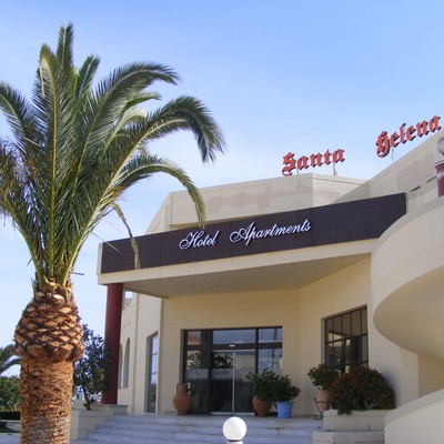 Entrance of Santa Helena Hotel Apartments in Crete, featuring a modern cream-colored building with potted plants, a palm tree, and clear blue skies.