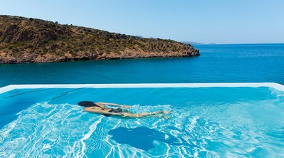 A serene infinity pool overlooking the turquoise waters of a secluded cove, with a guest relaxing against the backdrop of rugged hills and the endless blue sea, at the Daios Cove Luxury Resort A serene infinity pool overlooking the turquoise waters of a secluded cove, with a guest relaxing against the backdrop of rugged hills and the endless blue sea, at the Daios Cove Luxury Resort