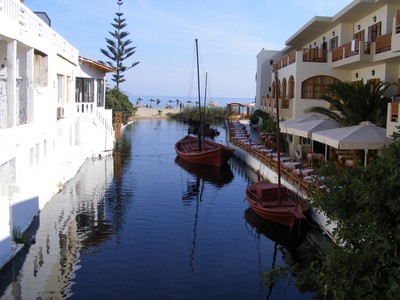 A charming canal scene in Kalyves village, Crete, with traditional wooden boats moored along the water, lined by whitewashed buildings and a cozy outdoor café. The canal opens up to the beach in the distance, creating a peaceful and picturesque setting that reflects the relaxed atmosphere of this coastal village. A charming canal scene in Kalyves village, Crete, with traditional wooden boats moored along the water, lined by whitewashed buildings and a cozy outdoor café. The canal opens up to the beach in the distance, creating a peaceful and picturesque setting that reflects the relaxed atmosphere of this coastal village.