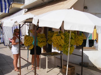 A small outdoor banana stall in Crete, shaded by white umbrellas, selling bunches of locally grown bananas. Two women are selecting fruit, while another arranges the display against the backdrop of a traditional Greek building. A small outdoor banana stall in Crete, shaded by white umbrellas, selling bunches of locally grown bananas. Two women are selecting fruit, while another arranges the display against the backdrop of a traditional Greek building.