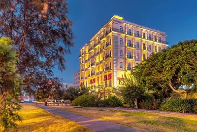 A beautifully illuminated multi-story image of the Megaron hotel in Herakion at dusk, featuring classic architecture with large windows and balconies. The hotel is surrounded by lush greenery, with a paved pathway leading through a park-like area in the foreground, under a clear evening sky.