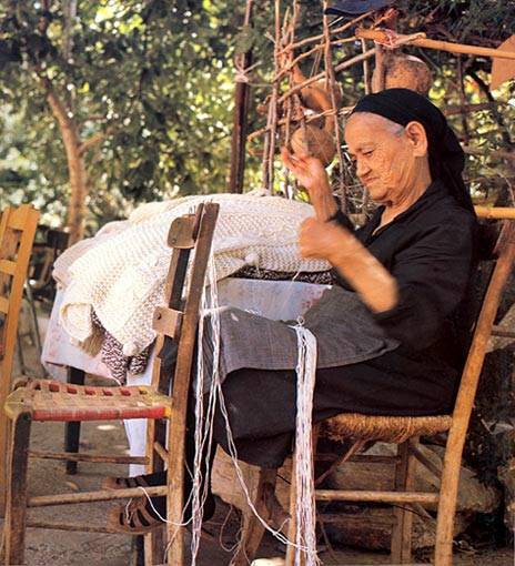 An elderly woman in traditional Cretan attire skillfully crafting lace by hand while seated outdoors, surrounded by wooden chairs and leafy trees.