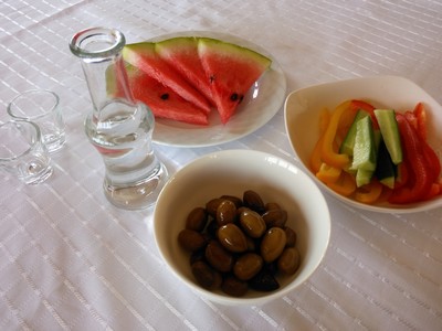 A traditional Cretan Raki serving with a clear glass carafe of Raki, two small shot glasses, and bowls of olives, sliced watermelon, and fresh vegetable sticks, set on a white tablecloth. A traditional Cretan Raki serving with a clear glass carafe of Raki, two small shot glasses, and bowls of olives, sliced watermelon, and fresh vegetable sticks, set on a white tablecloth.