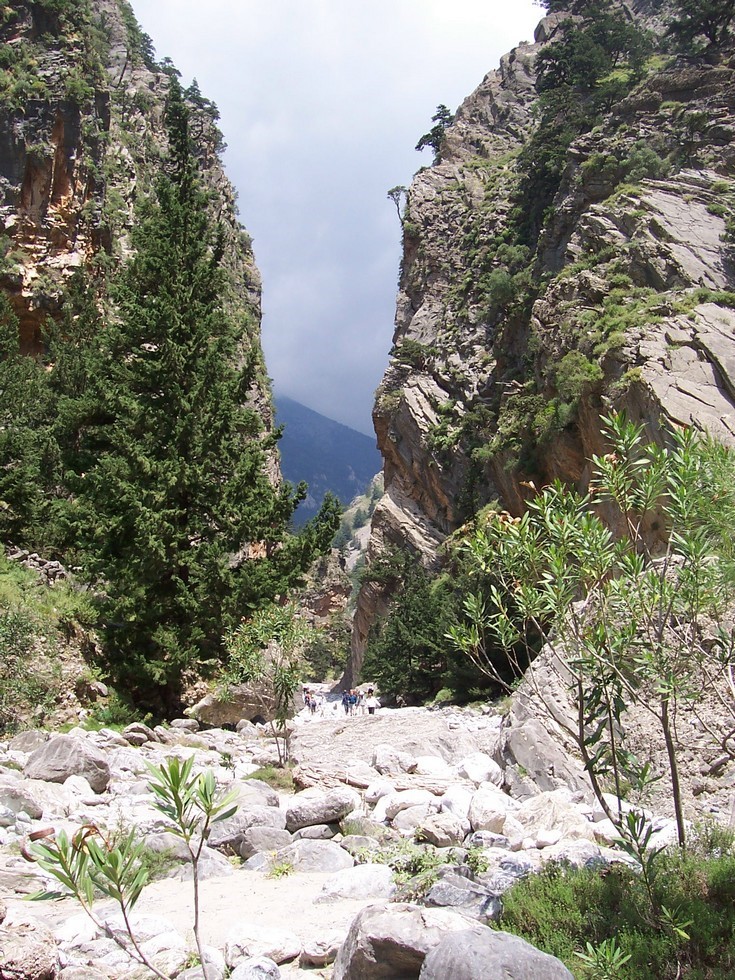 View of the Samaria Gorge in Crete, with towering rock formations, lush greenery, and hikers exploring the rocky trail below. View of the Samaria Gorge in Crete, with towering rock formations, lush greenery, and hikers exploring the rocky trail below.