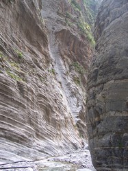 Narrow section of Samaria Gorge in Crete, Greece, showcasing towering, rugged limestone cliffs with distinct striations, a rocky path below, and patches of greenery clinging to the steep rock faces.