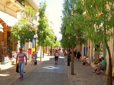 A lively shopping street in Agios Nikolaos, Crete, lined with trees, boutique shops, and cafés. People stroll along the pedestrian-friendly walkway, browsing souvenirs, fashion, and local goods under the warm Mediterranean sun.