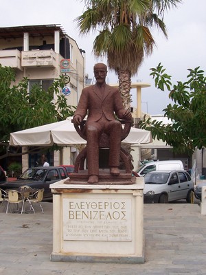 Statue of Eleftherios Venizelos in Kissamos, Crete, surrounded by greenery, a palm tree, and local buildings in the village square.