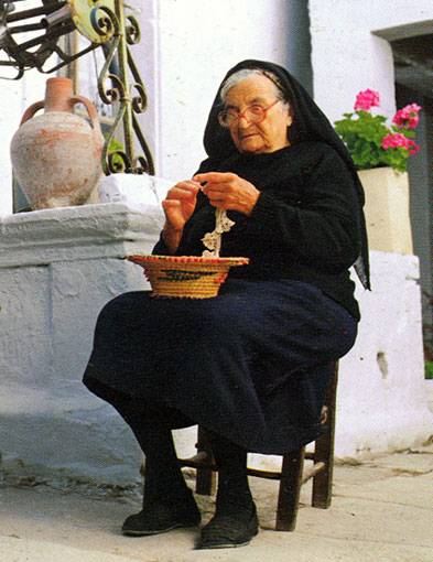 An elderly Cretan woman dressed in traditional black clothing, sitting on a wooden stool outside a whitewashed house, skillfully weaving lace in a peaceful village setting. An elderly Cretan woman dressed in traditional black clothing, sitting on a wooden stool outside a whitewashed house, skillfully weaving lace in a peaceful village setting.