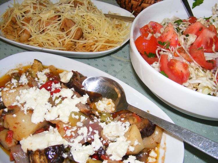 A close-up of a traditional Cretan meal featuring three dishes. In the foreground, there's a vegetable stew topped with crumbled feta cheese. To the left, a plate of pasta with grated cheese and small pieces of meat. On the right, a fresh salad with chopped tomatoes, cabbage, and parsley, lightly dressed. The dishes are served in white bowls on a light green tablecloth. A close-up of a traditional Cretan meal featuring three dishes. In the foreground, there's a vegetable stew topped with crumbled feta cheese. To the left, a plate of pasta with grated cheese and small pieces of meat. On the right, a fresh salad with chopped tomatoes, cabbage, and parsley, lightly dressed. The dishes are served in white bowls on a light green tablecloth.